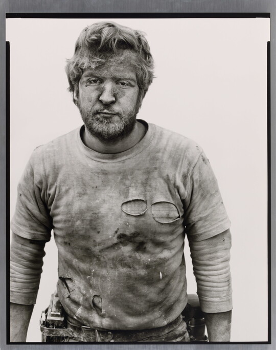 A black-and-white portrait photograph of a White man with messy hair and a beard wearing a dirty, ripped shirt.