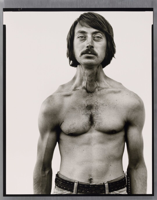 A black-and-white portrait photograph of a shirtless White man, with dark hair and a mustache, and thin but with muscled shoulders and arms.