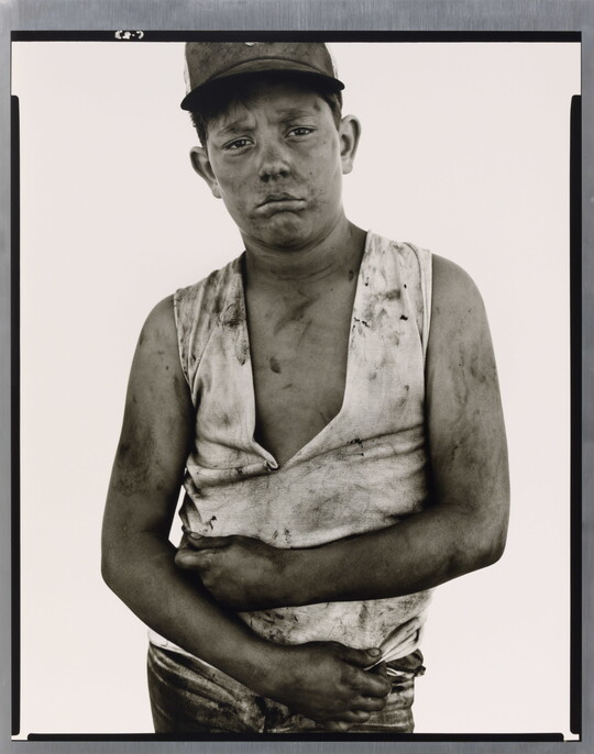 A black-and-white portrait photograph of a dirty young man wearing a baseball hat, a dirty ripped shirt, and holding his arms across his torso.