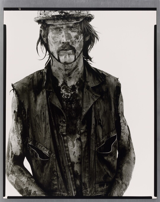 A black-and-white portrait photograph of a White man covered in dirt and oil wearing a vest and hard hat.