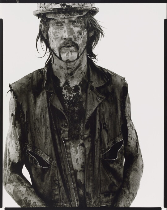 A black-and-white portrait photograph of a White man covered in dirt and oil wearing a vest and hard hat.