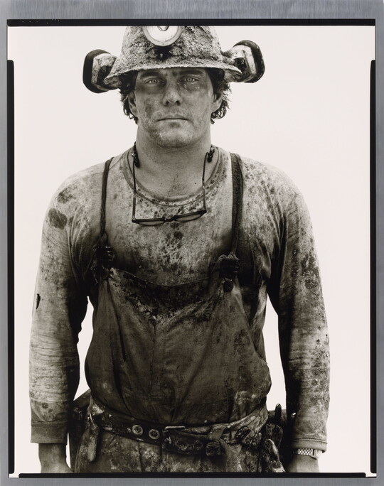 A black-and-white portrait photograph of a young White man covered in dirt wearing a hard hat and a shirt under coveralls.