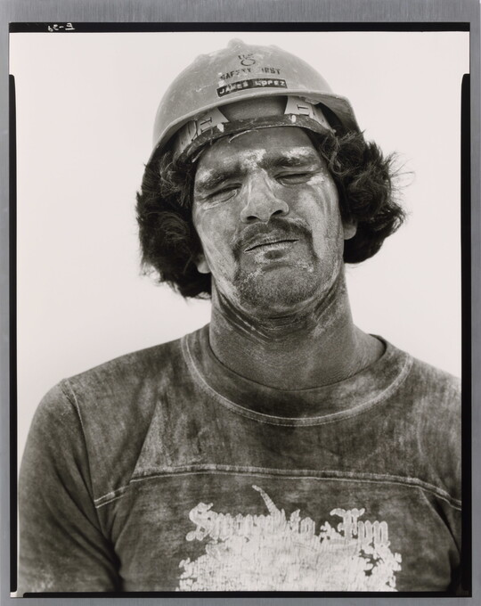 A black-and-white portrait photograph of a man wearing a hard hat, his face and neck smeared with white dust, and dirty t-shirt.