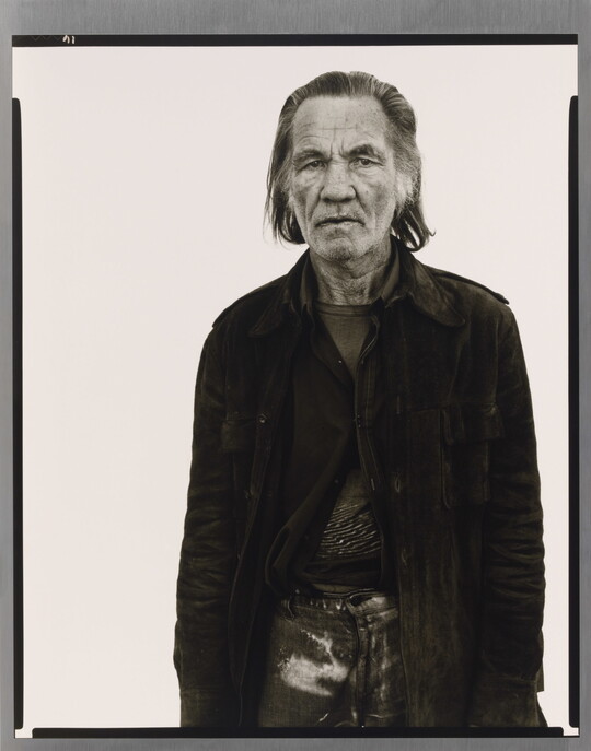 A black-and-white portrait photograph of an older White man with long hair wearing a partially-buttoned jacket over his clothing.