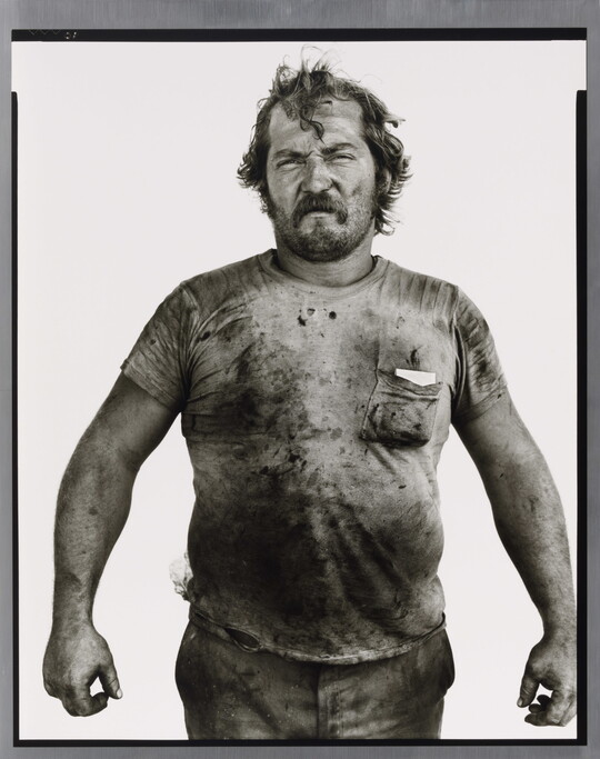 A black-and-white portrait photograph of a White man with messy hair and a beard wearing a dirty t-shirt and holding his arms out from his sides.