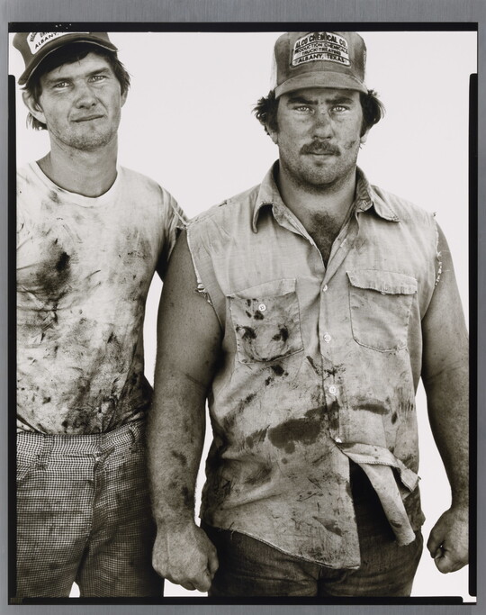 A black-and-white portrait photograph of two White men wearing dirty clothes and baseball caps standing next to each other.