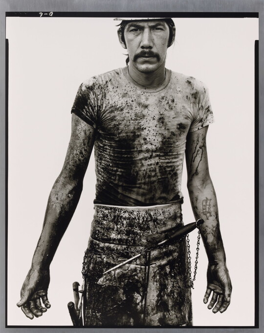 A black-and-white portrait photograph of a White man with a mustache wearing a dirty t-shirt and apron holding with different tools on it.