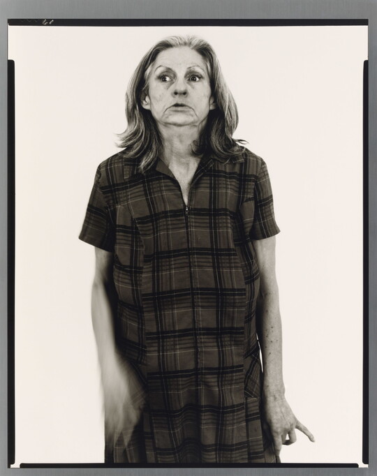 A black-and-white portrait photograph of an older White woman with medium-length gray hair, head held back slightly, wearing a house dress.
