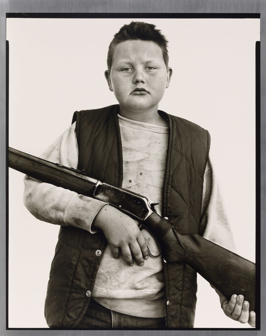 A black-and-white portrait photograph of a White boy with close cropped hair, wearing a quilted vest over a sweatshirt, and holding a rifle across his torso.