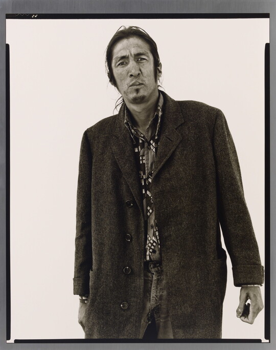 A black-and-white portrait photograph of a medium-skinned man with long hair pulled back, a goatee, and wearing a long wool coat.