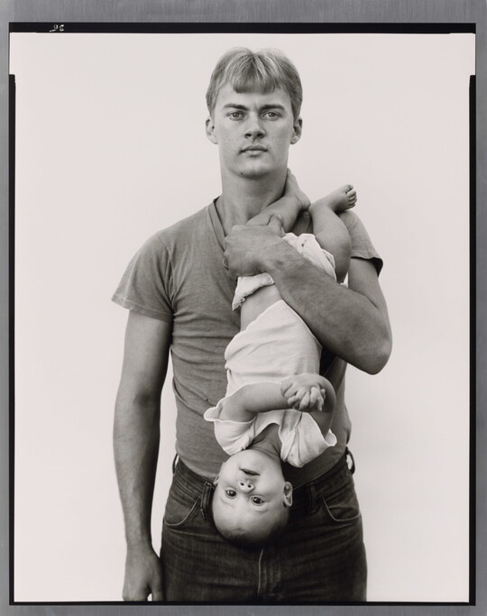 A black-and-white portrait photograph of a White man with light hair holding a baby upside down in front of his chest.