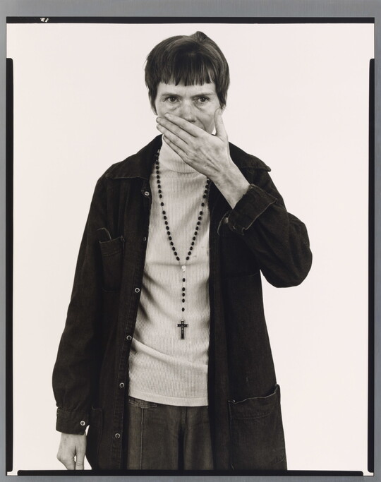 A black-and-white portrait photograph of a White woman with short, dark hair wearing a dark jacket and rosary beads around her neck holding one hand in front of her mouth and nose.