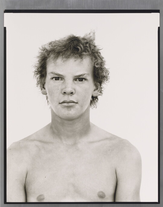 A black-and-white portrait photograph of a shirtless White teenage boy with messy hair.