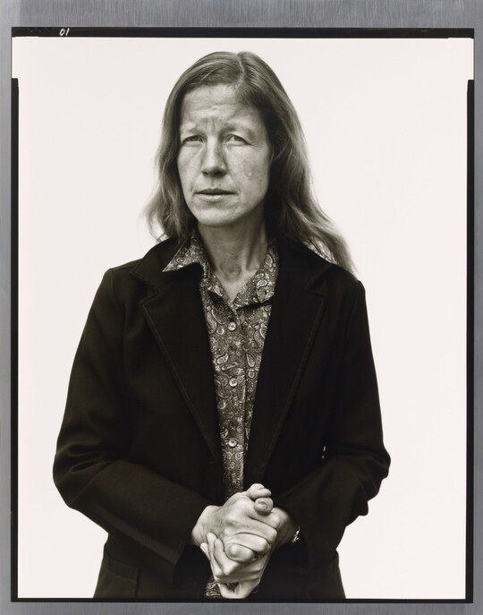 A black-and-white portrait photograph of a White woman with long hair wearing a button-down blouse under a suit jacket holding her hands clasped in front of her.