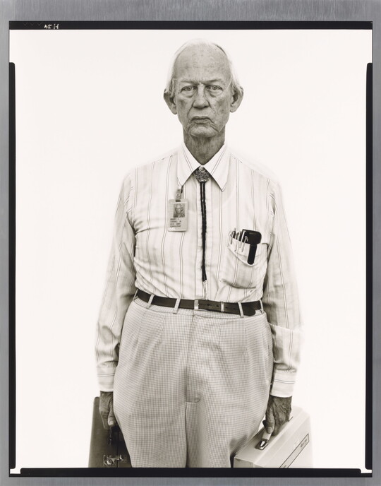 A black-and-white portrait photograph of an older, mostly bald White man wearing a collared shirt, a bolo tie, and work badge attached to his collar, holding two briefcases.