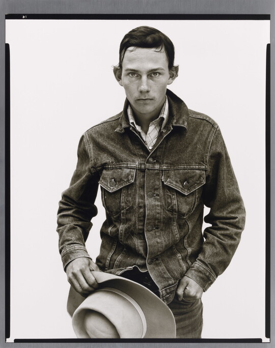 A black-and-white portrait photograph of a young White man with short dark hair wearing a denim jacket and holding a cowboy hat in front of him.