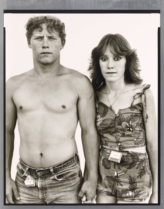 A black-and-white portrait photograph of a young, shirtless White man holding hands with a young White woman wearing a tank top and shorts.
