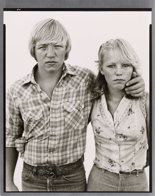A black-and-white portrait photograph of a White teenage boy wearing a plaid button-down shirt, his arm around the shoulders of a White teenage girl wearing a sleeveless shirt.
