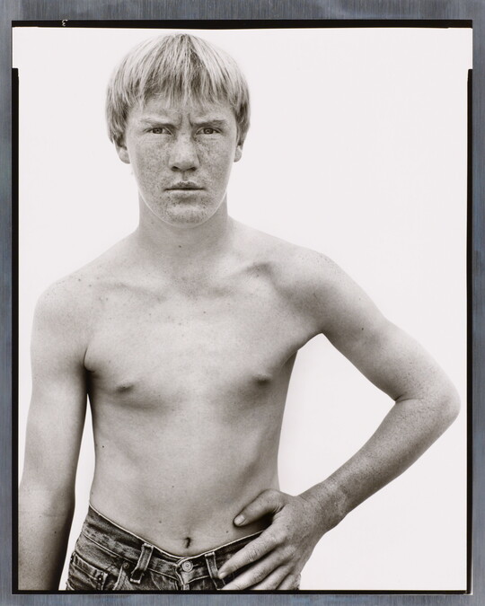 A black-and-white photograph of a shirtless White boy with light hair and a freckled face standing with one hand on his hip.