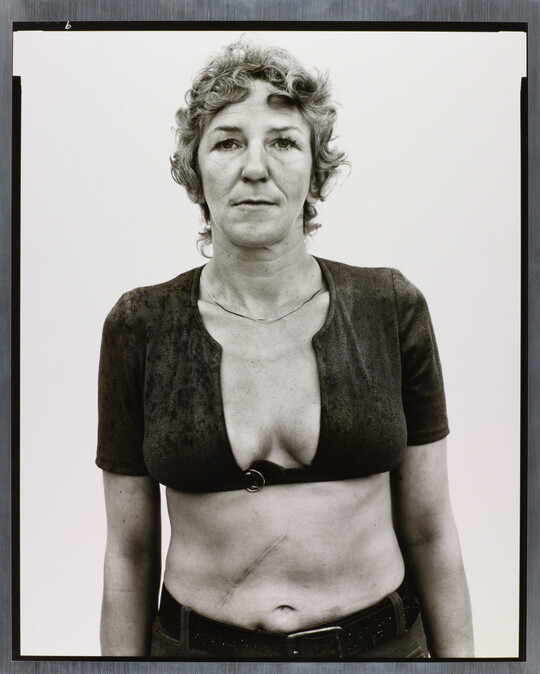 A black-and-white portrait photograph of a White woman with short, curly hair wearing a cropped shirt with a very low neckline.