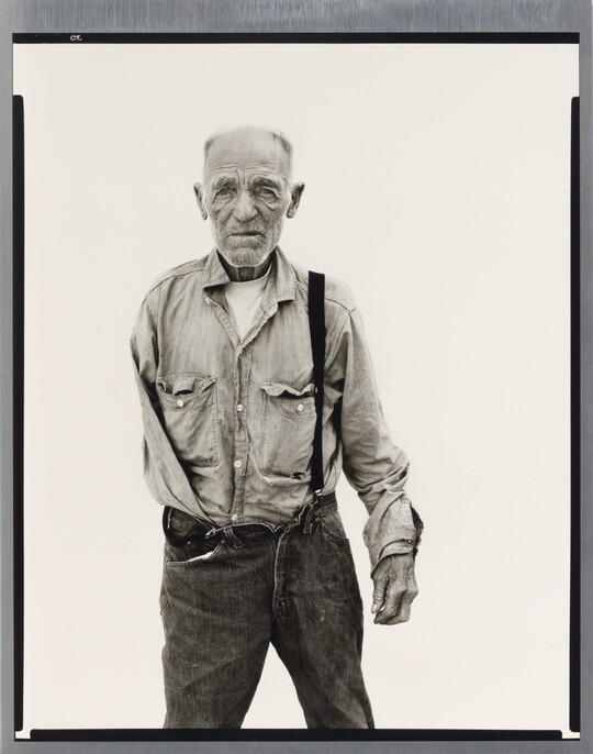 A black-and-white portrait photograph of an older, balding White man with one arm wearing a button-down shirt and jeans held up with one suspender.