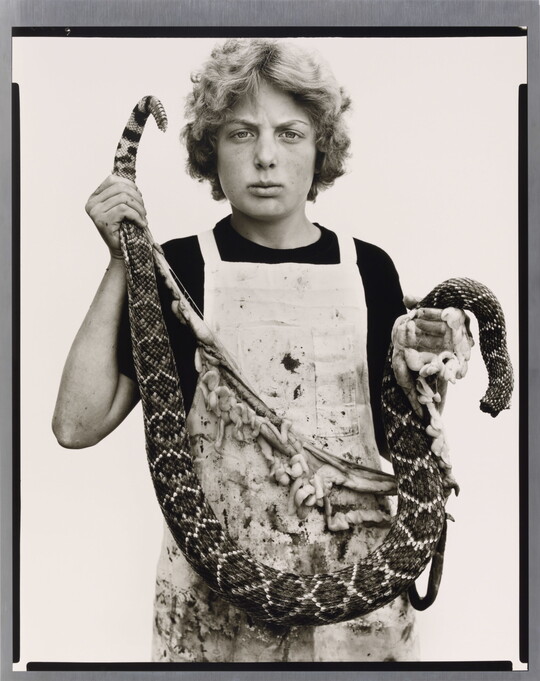A black-and-white portrait photograph of a White teenage boy with curly hair wearing a dirty apron and holding a gutted snake in front of him.