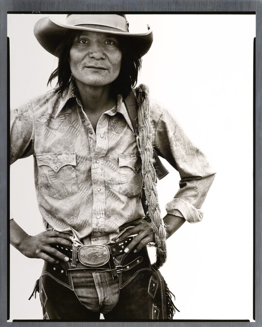A black-and-white portrait photograph of a Native American man with long hair wearing a cowboy hat, button-down shirt, jeans, and chaps, standing with hands on hips.