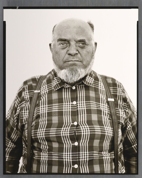 A black-and-white portrait photograph of an older, balding White man with a gray beard wearing a plaid button-down shirt and suspenders.