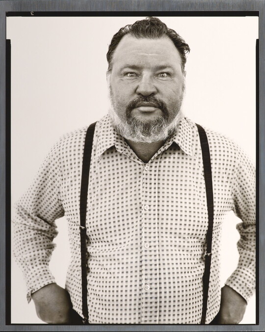 A black-and-white portrait photograph of a middle-aged White man with a receding hairline and salt-and-pepper beard wearing a checked button-down shirt and suspenders.