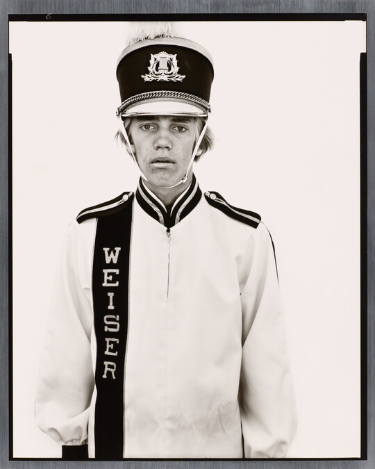 A black-and-white portrait photograph of a White teenage boy wearing an oversized, white, marching-band hat and uniform.