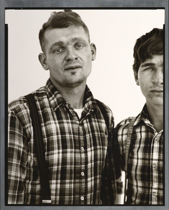A black-and-white portrait photograph of two White men, both in similar plaid shirts and suspenders, standing shoulder to shoulder.