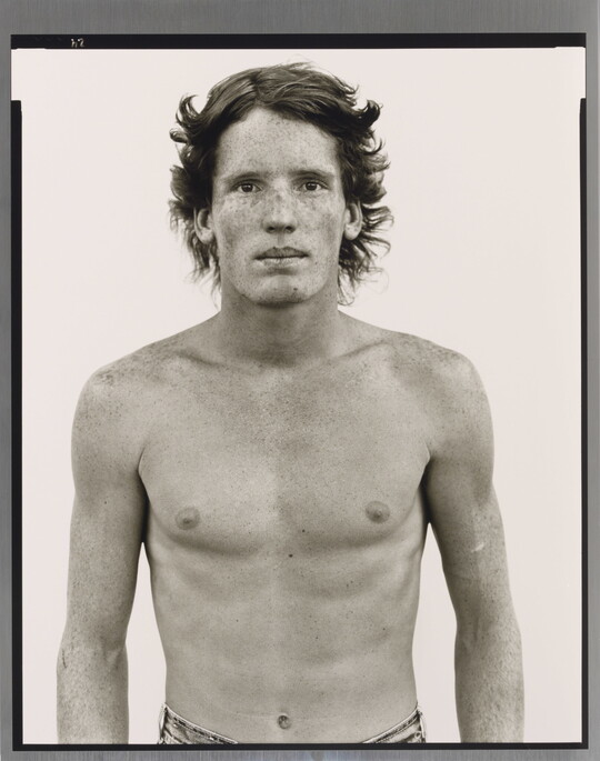A black-and-white portrait photograph of a shirtless young White man with freckles across his face and shoulders.