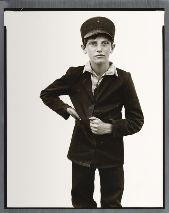 A black-and-white portrait photograph of a White boy wearing a Breton cap, a dark button-up jacket with one hand on his waist and the other tucked in between the buttons.