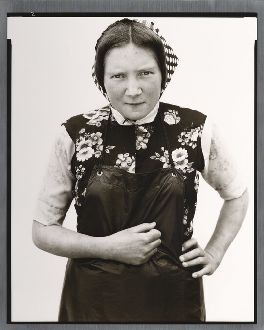A black-and-white portrait photograph of young White woman wearing a checked kerchief, flowered vest, and dark apron.