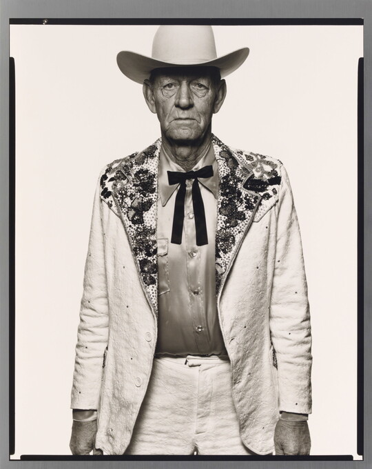 A black-and-white portrait photograph of an older White man wearing a white cowboy hat, a white suit jacket with rhinestones on the collar, a satin shirt, and a bow tie.