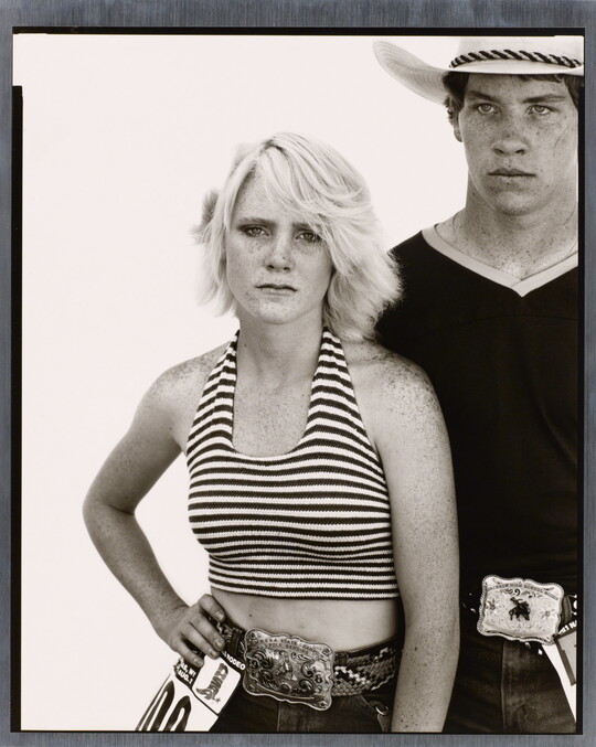A black-and-white portrait photograph of a young White woman wearing a striped halter top, one hand on her hip, and a young White man wearing a cowboy hat and V-neck t-shirt, both wearing large buckles on their belts.