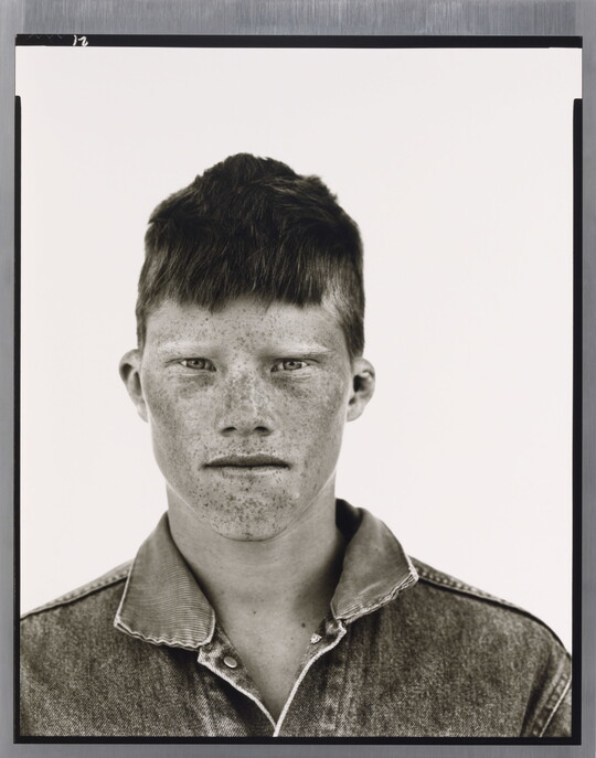 A black-and-white portrait photograph of a White teenage boy, face covered in freckles, short cropped hair, wearing a denim jacket.