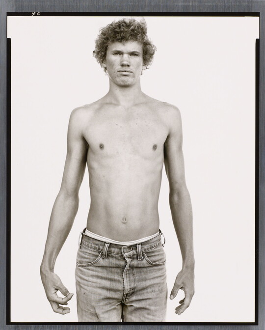 A black-and-white portrait photograph of a shirtless, curly-haired, teenage, White boy in jeans holding his hands by his sides.
