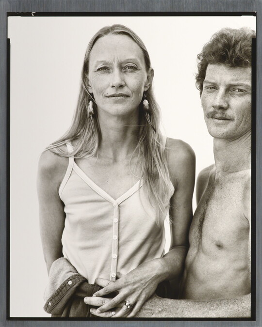 A black-and-white portrait photograph of a thin White woman with long hair wearing camisole as a shirtless White man with curly hair and a moustache holds her waist.