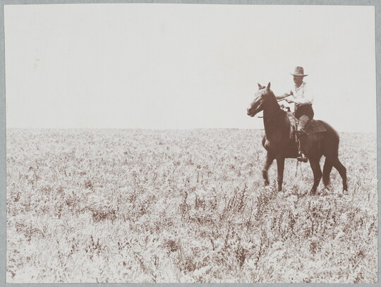 A black-and-white photograph of a man wearing a cowboy hat riding a horse in an open field.