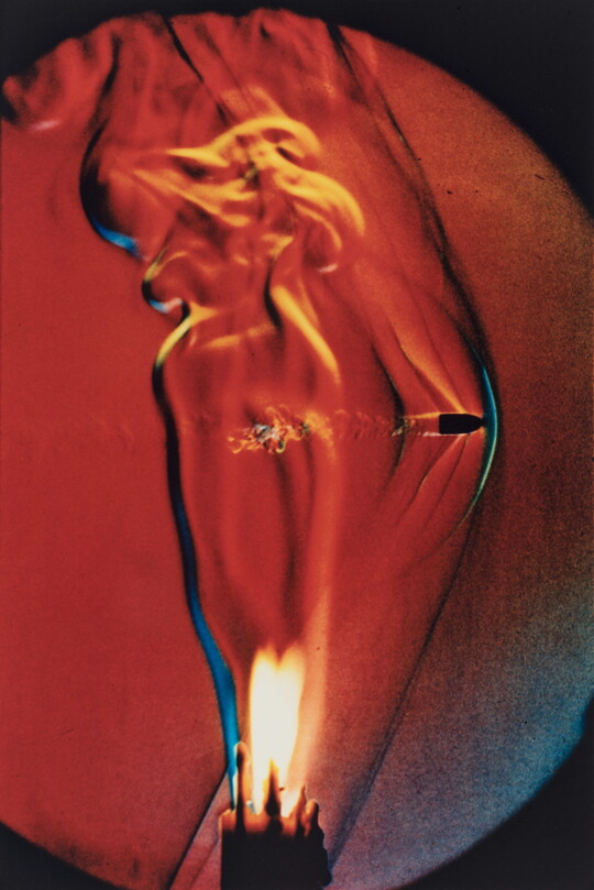 A color photograph of a lit candle with a bullet going through the flame.