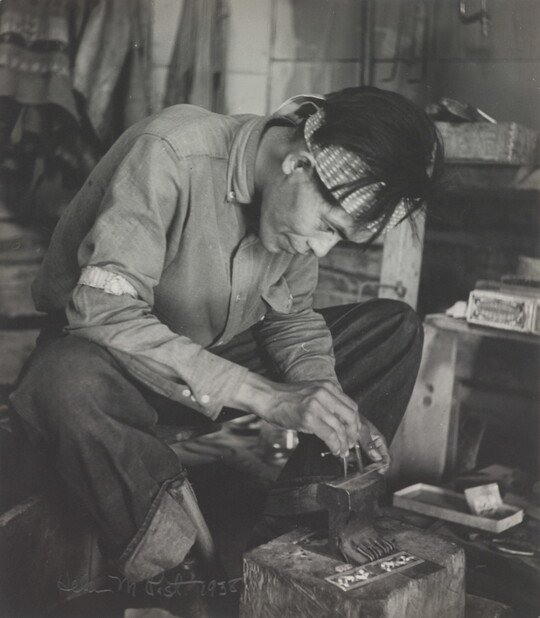 A black-and-white photograph of a seated Native American man bent over an anvil in a workshop.