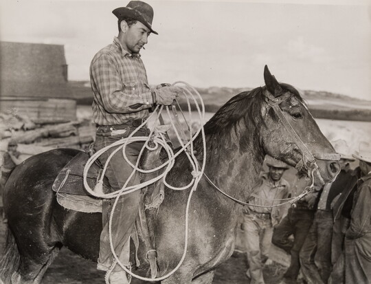 A black-and-white photograph of a light-skinned man in a brimmed hat holding a lasso on horseback.