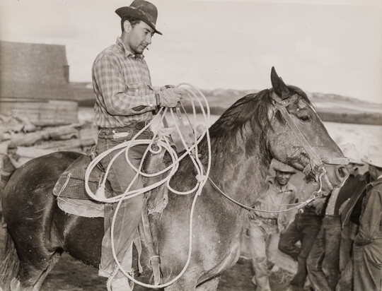 A black-and-white photograph of a light-skinned man in a brimmed hat holding a lasso on horseback.