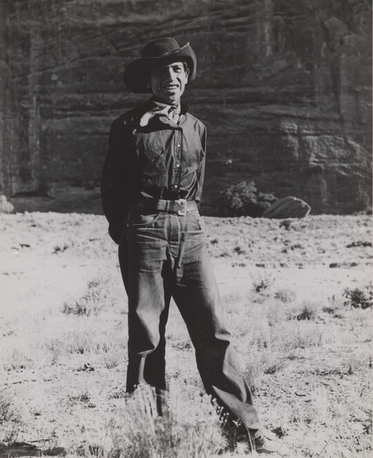 A black-and-white photograph of a man wearing a cowboy hat and neckerchief posing in front of what looks like the face of a cliff.