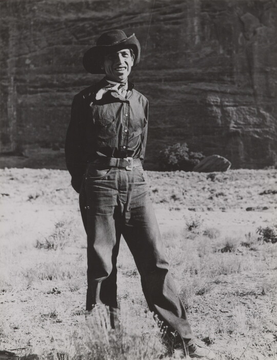 A black-and-white photograph of a man wearing a cowboy hat and neckerchief posing in front of what looks like the face of a cliff.