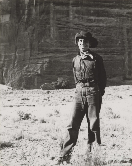 A black-and-white photograph of a man wearing a cowboy hat and neckerchief posing in front of what looks like the face of a cliff.