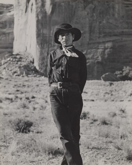 A black-and-white photograph of a man wearing a cowboy hat and neckerchief posing in front of what looks like the face of a cliff.