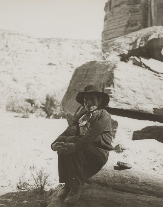 A black-and-white photograph of a man wearing a cowboy hat and neckerchief sitting on a rock in the shadows of a cliff face.