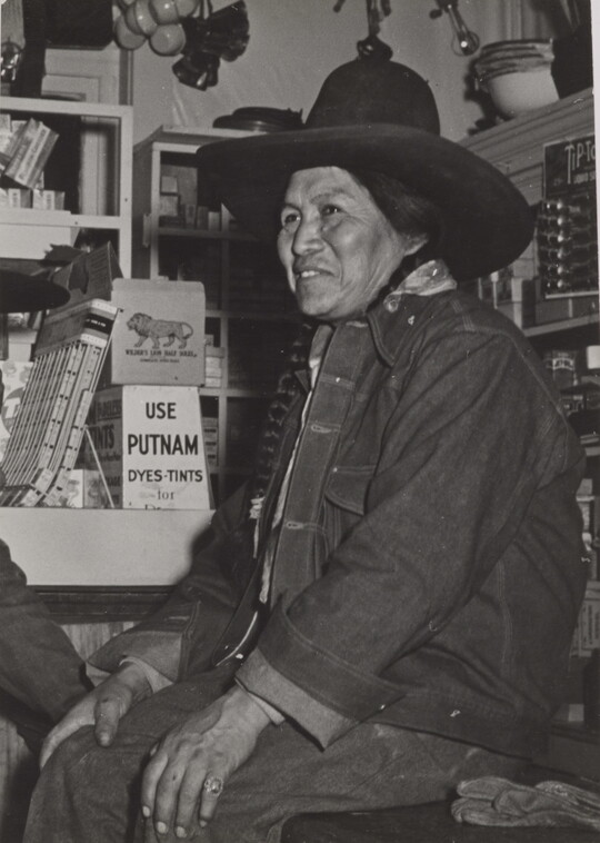 A black-and-white photograph of a Native American person wearing a jean jacket and cowboy hat sitting on a counter in a general store.